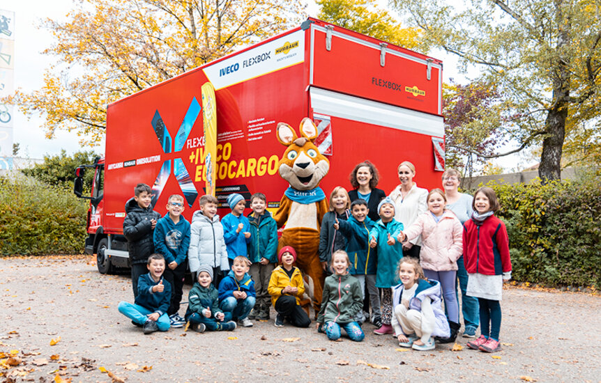Grundschulklasse steht mit Maskottchen Blicki, Frau Humbaur und Frau Friedrichs vor einem LKW auf dem Schulhof | © Humbaur GmbH