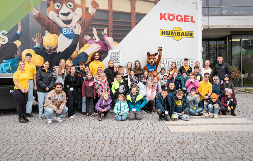 Kinder und das Blicki-Maskottchen stehen vor einem LKW der mit den Logos von Kögel und Humbaur bedruckt ist | © Humbaur GmbH
