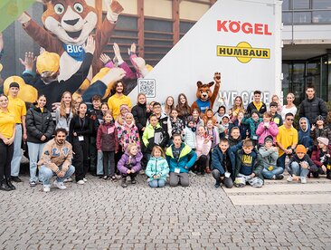 Kinder und das Blicki-Maskottchen stehen vor einem LKW der mit den Logos von Kögel und Humbaur bedruckt ist | © Humbaur GmbH