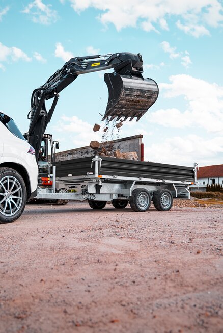 Mit einem Bagger werden Baumaterialien in einen Kastenanhänger geschüttet. Der Anhänger ist an ein weißes Auto angekoppelt. | © Humbaur GmbH